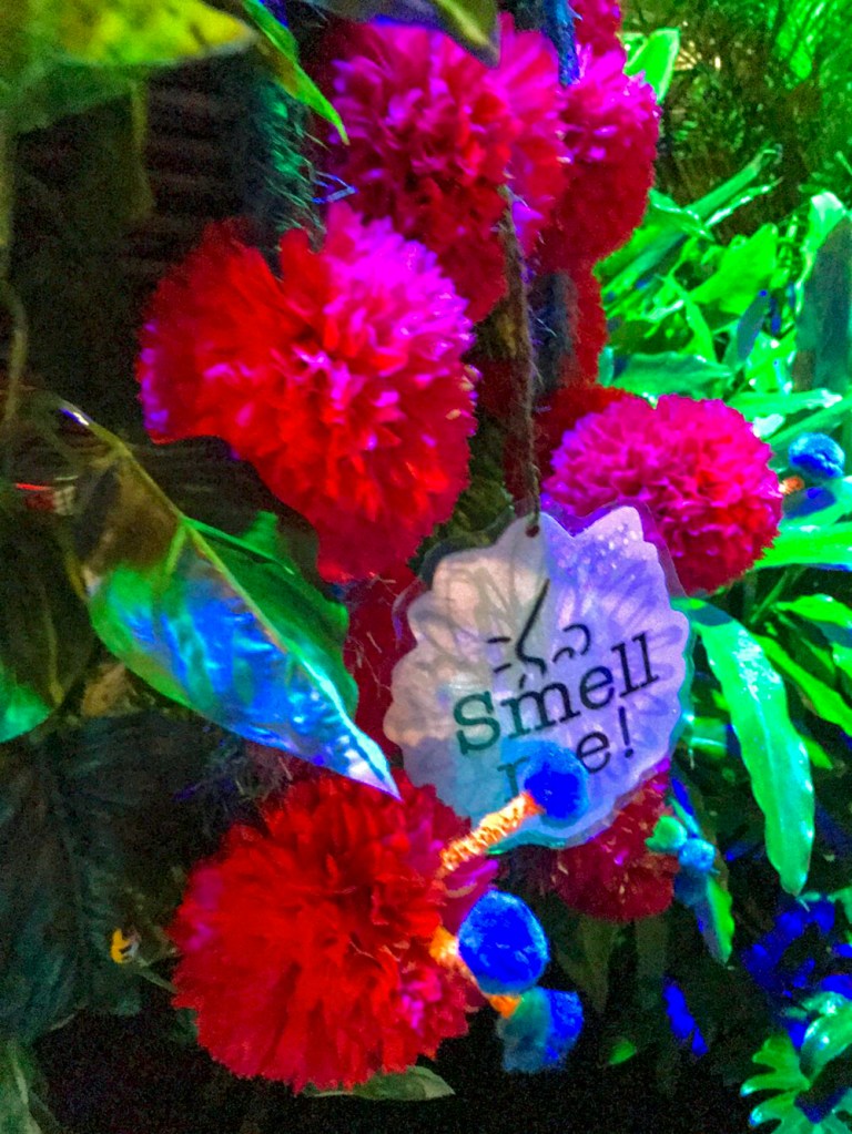 In the midst of flowers, a single sign that says "Smell me!" with the drawing of a nose.